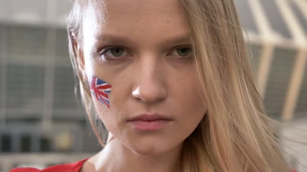 Portret van een jonge blonde meisje voetbal fan van Engeland op zoek naar de camera, een ernstig gezicht, een stadion in de achtergrond 50 fps — Stockvideo