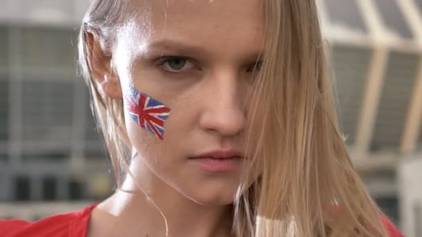 Retrato de una joven chica rubia fanática del fútbol de Inglaterra mirando a la cámara, cara seria, estadio en el fondo 50 fps — Vídeos de Stock