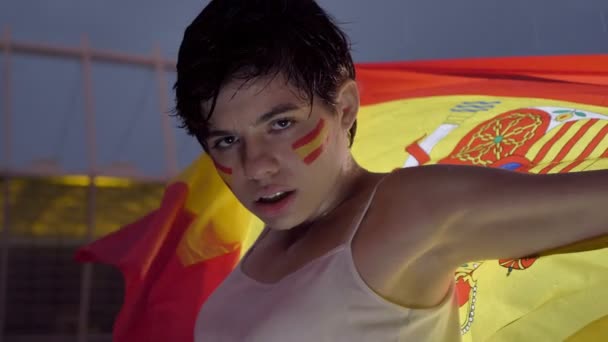 Retrato de uma jovem morena fã de futebol na Espanha, na chuva, olhando para a câmera, rosto sério, segurando uma bandeira atrás dela beck, estádio à noite no fundo 50 fps — Vídeo de Stock