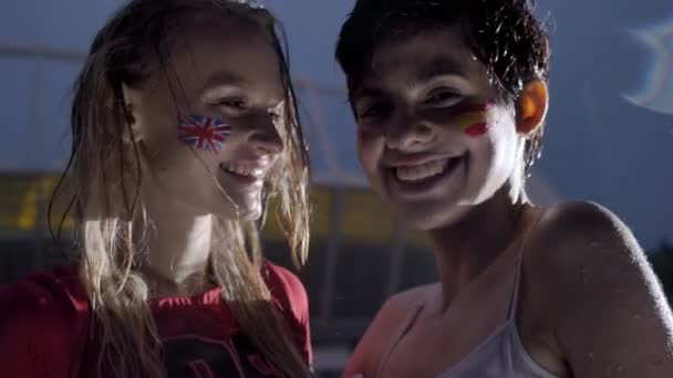 Δύο νεαρά κορίτσια, οπαδοί του ποδοσφαίρου σε βροχή, Αγγλία και Ισπανία, χαμογελώντας, γελώντας, στάδιο στο βάθος 50 fps — Αρχείο Βίντεο