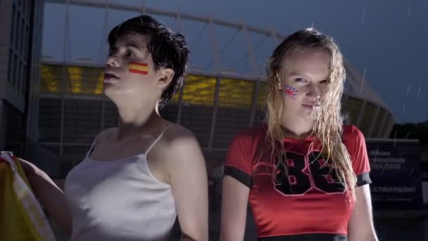 Liga dos Campeões, duas meninas fãs de futebol na chuva, confronto, Espanha e Inglaterra, estádio em segundo plano 60 fps — Vídeo de Stock