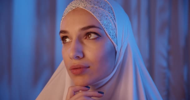 Retrato de una chica caucásica en una luz de neón hijab blanco mirando a la cámara pensando en maquillaje nocturno serio fondo azul — Vídeos de Stock