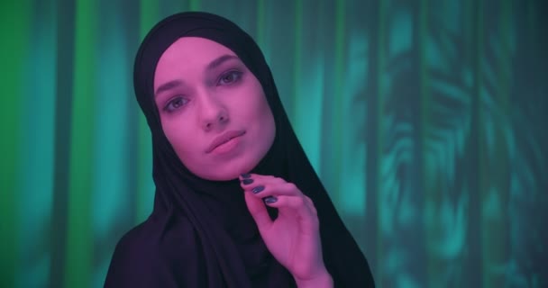 Beautiful confident girl in black hijab neon night green tropical background shadow image make-up purple color profile view turn rotates touches face