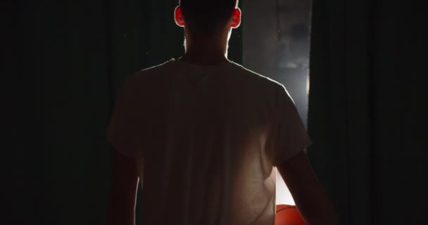 Jonge blanke basketbalspeler klaar om te gaan backstage duisternis silhouet straal van licht vertrouwen bal training verblinding achtergrond sportschool — Stockvideo