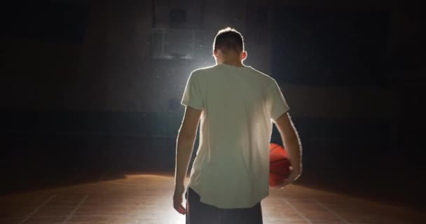 Mladý kavkazský basketbalista se chystá jít do zákulisí temnota silueta paprsek světla sebevědomí hraje s míčkem oslnění pozadí parkety — Stock video