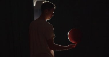 Yakın plan beyaz tenli basketbolcu bir topla oynamaya hazırlanıyor. Eğitimler parmağında döner sahne arkasındaki karanlık gölge ışık huzmesi