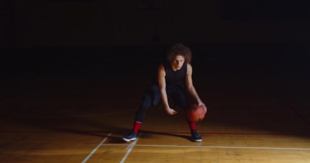 Krullend blank basketbal speler dribbelen bal camera professionele duisternis straal van licht — Stockvideo