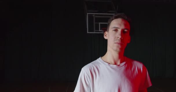 Primer plano caucásico joven jugador de baloncesto retrato rojo haz de luz negro oscuro cesta fondo — Vídeos de Stock