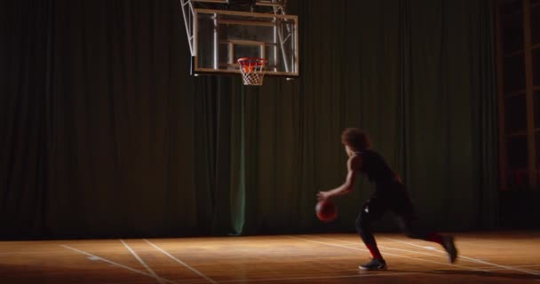 Joven jugador de baloncesto rizado juega parquet lanza la pelota en la oscuridad canasta noche — Vídeo de stock