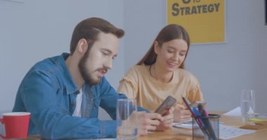 Güneşli bir ofisteki genç meslektaşlar gülümseyen gülüşler gösteren akıllı telefonlar kullanır.