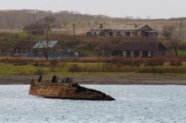 Harap olmuş ahşap bir gemi, ahşap evleri olan bir adanın arka planına karşı deniz suyundan sarkıyor.