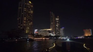 2019 yazında Bangkok, Tayland 'da Bangkok' un ana nehri Chhao Praia boyunca bir turist teknesinde gece yürüyüşü. Turistlerle dolu bir tekne yüksek binaların ve parlak alışveriş merkezlerinin önünden yüzüyor..
