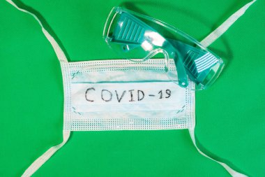 Üzerinde siyah Covid-19 yazan steril tıbbi bir maske ve yeşil bir arka planda duran güvenlik gözlükleri..