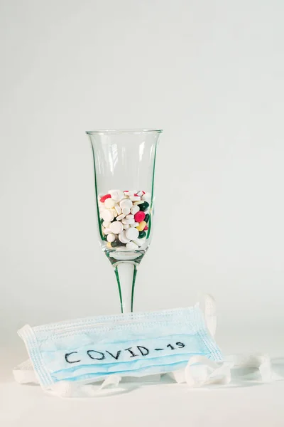 Coronavirus Covid -19. Champagne glass half filled with different pills is on a white background. Sterile medical mask with the inscription Covid -19.