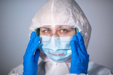 COVID-19. 2019 Novel Coronavirus (2019-nCoV) konsepti. Beyaz koruyucu önlüklü ve tıbbi maskeli genç bir doktor Coronovirus yazısıyla güvenlik gözlüklerini elleriyle ayarlıyor.