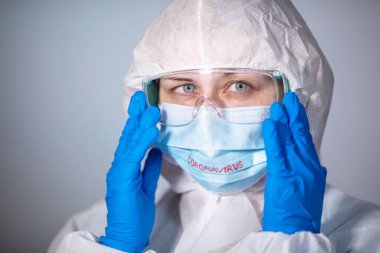 COVID-19. 2019 Novel Coronavirus (2019-nCoV) konsepti. Beyaz koruyucu önlüklü ve tıbbi maskeli genç bir doktor Coronovirus yazısıyla güvenlik gözlüklerini elleriyle ayarlıyor.