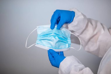 COVID-19. Yığının üzerinde yüz maskeleri var. Salgın geçmişi. Yakın plan. Koruyucu veba önleme giysisi giymiş bir tıp çalışanı plastik tek kullanımlık eldivenli bir maske tutuyor.