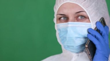 COVID-19. 2019 Novel Coronavirus 2019-nCoV konsepti. Genç bayan doktor telefonda koruyucu bir maske ve yeşil bir arka planda lastik eldivenle beyaz sisli bir ceketle konuşuyor.