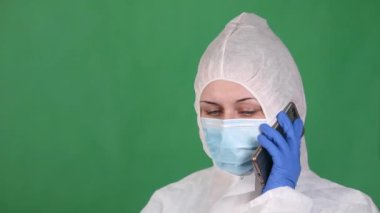 COVID-19. 2019 Novel Coronavirus 2019-nCoV konsepti. Genç bayan doktor telefonda koruyucu bir maske ve yeşil bir arka planda lastik eldivenle beyaz sisli bir ceketle konuşuyor.