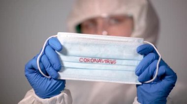 COVID-19. 2019 Novel Coronavirus (2019-nCoV) konsepti. Beyaz koruyucu veba önlüklü genç bir doktor elinde Coronavirus yazısıyla tıbbi bir maske tutuyor.