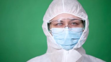 COVID-19. 2019 Novel Coronavirus (2019-nCoV) konsepti. Genç kız doktor. Elinde veba önlüğü, tıbbi maske ve yeşil arka planda koruyucu şeffaf gözlükler olan kameraya bak..