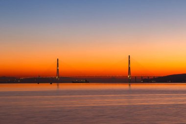 Deniz kenti Vladivostok 'ta şafak vakti. Vladivostok 'taki Russky Adası' na uzanan Vostochny Boğazı 'ndaki Rus köprüsünde, sakin deniz sırasında, altın bir şafağın arka planına karşı kablo destekli Rus köprüsü..