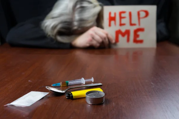 Drogově závislý u stolu drží znak m s žádostí o pomoc, před ním stříkačky léků. — Stock fotografie