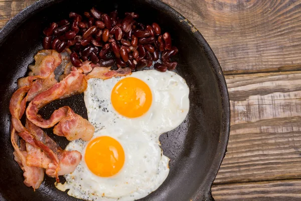 Bacon, uova e fagioli. Uovo salato e cosparso di pepe nero. Colazione inglese. Pancetta alla griglia, due uova e fagioli in padella sul tavolo di legno — Foto Stock