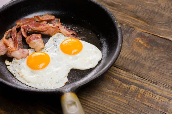 Bacon i jaj. Solone jaja i posypane pieprzem. Śniadanie angielskie. Boczek z grilla, dwa jaja na patelni na drewnianym stole. — Zdjęcie stockowe