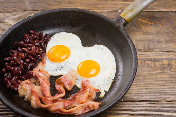 Speck, Ei und Bohne. gesalzenes Ei und mit schwarzem Pfeffer bestreut. Englisches Frühstück. gegrillter Speck, zwei Eier und Bohnen in Pfanne auf Holztisch — Stockfoto