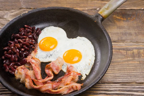 Slanina, vejce a fazole. Osolené vejce a počítat s černým pepřem. Anglická snídaně. Grilovaná slanina, dvě vejce a fazole v pánvi na dřevěný stůl — Stock fotografie