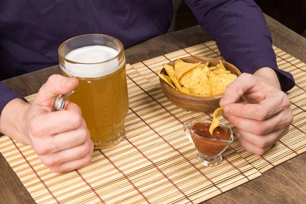 Mesa de madera manos masculinas sosteniendo una taza con cerveza y nachos sumergidos en la salsa. Copiar pasta — Foto de Stock