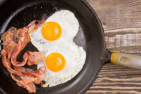 Bacon i jaj. Solone jaja i posypane pieprzem. Śniadanie angielskie. Boczek z grilla, dwa jaja na patelni na drewnianym stole. — Zdjęcie stockowe