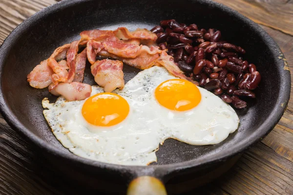 Bacon, uova e fagioli. Uovo salato e cosparso di pepe nero. Colazione inglese. Pancetta alla griglia, due uova e fagioli in padella sul tavolo di legno — Foto Stock