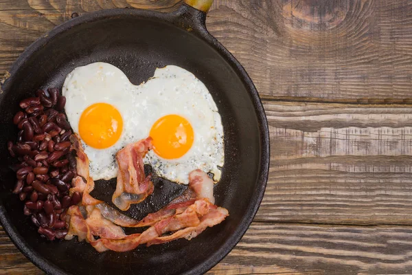 Bacon, uova e fagioli. Uovo salato e cosparso di pepe nero. Colazione inglese. Pancetta alla griglia, due uova e fagioli in padella sul tavolo di legno — Foto Stock