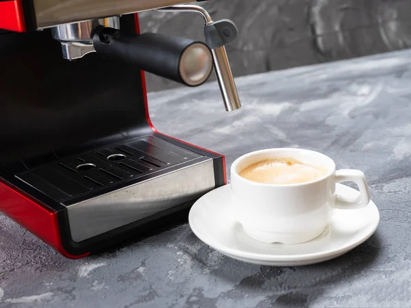 Uma xícara de café em um pires fica ao lado da cafeteira em uma bancada de concreto cinza . — Fotografia de Stock