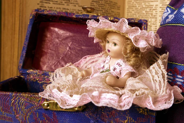 Uma boneca velha em um vestido openwork senta-se em um peito contra o pano de fundo de um livro velho. Foto vintage . — Fotografia de Stock