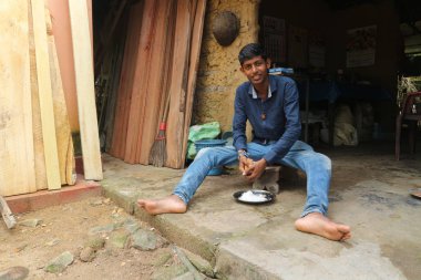  Sri Lanka, Berathuduwa - 2 Aralık 2019: Genç bir Sri Lankalı bir adam bir köy evinin kapısında oturur ve geleneksel otantik rende hindistan cevizi tanelerini ovuşturur.