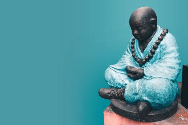 Mavi elbiseli bir Budist keşiş, kollarını kavuşturup meditasyona dalmış bir nilüfer pozisyonunda oturur. Koyu turkuaz arka planda taştan yapılmış Buda heykeli..