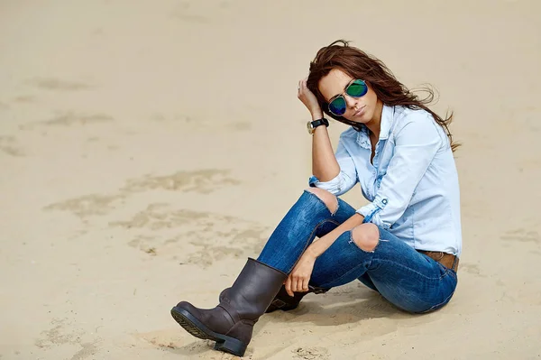 Kobieta w okularach i dżinsach siedzi na piasku - outdoo — Zdjęcie stockowe