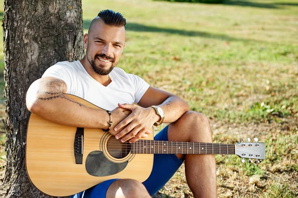 Красивый улыбающийся мужчина с акустической гитарой в летнем парке — стоковое фото