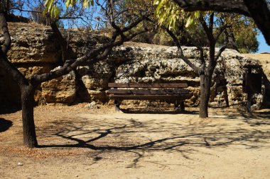 Önünde ağaçların gölgelerinin olduğu bir bank, antik bir duvarın arka planına karşı. Valle dei Templi, Agrigento, Sicilya, İtalya
