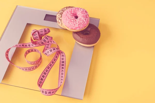 Delicious pink and chocolate donuts, a scale and a tailors meter on yellow background. Concept of diet and sweet temptation. — Stock Photo, Image