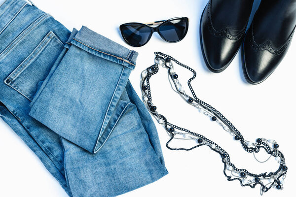 Overhead view of womans casual outfit on white background - glasses, blue jeans, necklace and leather chelsea boots. Flat lay, top view, copy space. Trendy, minimal hipster look.