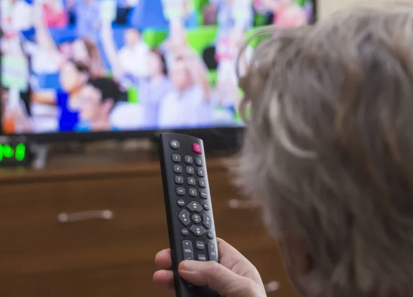 Arkadan bakıldığında, beyaz saçlı orta yaşlı bir kadın uzaktan kumandayla televizyonu kontrol ediyor.