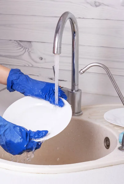 En kvinna i blå handskar tvättar en vit tallrik under rinnande vatten — Stockfoto