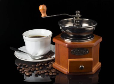 Kahve değirmeni ve bir fincan kahve ve kahve çekirdekleri siyah arka planda dağılmış.