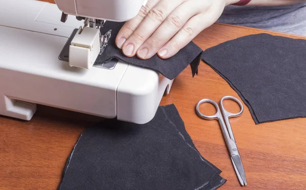 Bir kadın, karantina sırasında dışarıda takmak için dikiş makinesine siyah koruyucu bir maske dikiyor..