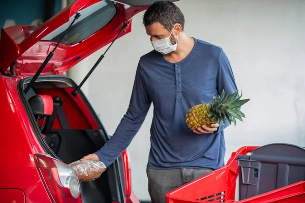 Młody mężczyzna nosi maskę medyczną przeciw wirusowi korony covid-19 na parkingu. Klient ładuje samochód z jedzeniem na parkingu supermarketu. Braki w zaopatrzeniu. Zakup i składowanie w panice. — Zdjęcie stockowe