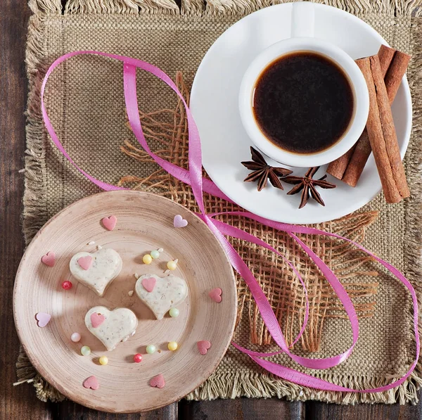 Milk sesame sweets in the form of heart and a cup of fragrant coffee with spices on an old wooden background.